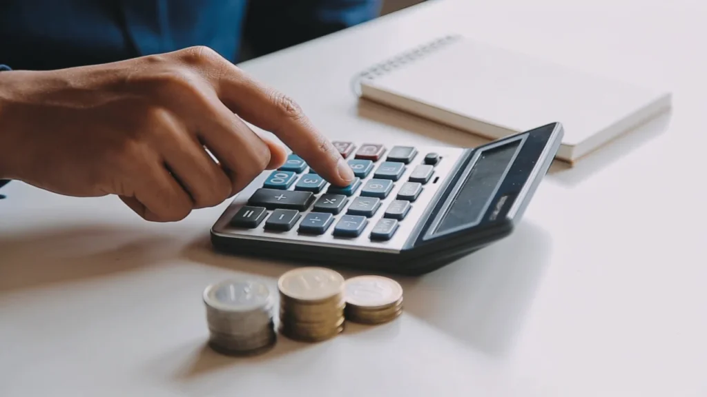 A hand pressing the keys of a desktop calculator beside a notebook and stacks of coins on a clean desk, representing calculating a financial independence number